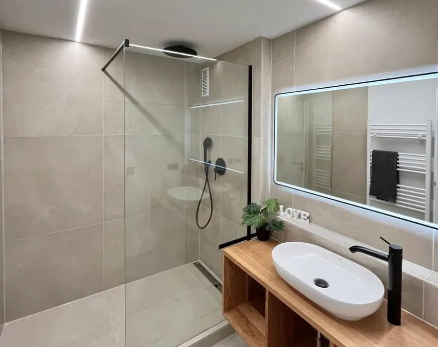 Modernes Badezimmer mit hellen Fliesen, einer begehbaren Dusche mit Glaswand, schwarzer Armatur und ovalem Waschbecken auf Holzarbeitsplatte.