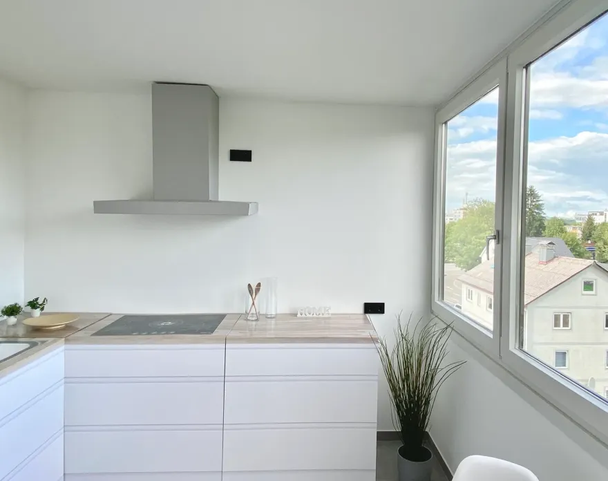 Moderne helle Küche mit weißen Schränken, Holzarbeitsplatte, Kochfeld und großer Fensterfront mit Blick auf Nachbarhäuser.