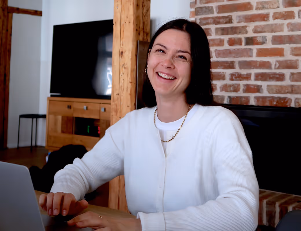 Lächelnde Frau mit dunklen Haaren, die einen weißen Pullover und eine goldene Halskette trägt, sitzt an einem Tisch mit einem Laptop in einem Raum mit Backsteinwand und Holzmöbeln.