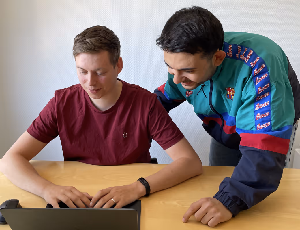 Zwei junge Männer an einem Schreibtisch schauen auf einen Laptop; einer sitzt und tippt, der andere steht und lehnt sich zum Beobachten herüber.