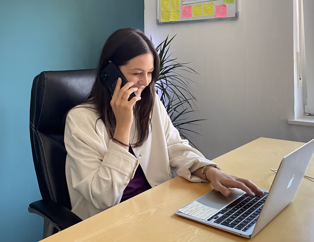 Lächelnde Frau, die am Telefon spricht, während sie auf einem Laptop an einem Holztisch in einem Büro tippt.