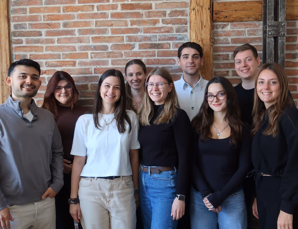 Lächelnde Gruppe von neun unterschiedlichen jungen Erwachsenen, die eng zusammen vor einer Backsteinwand stehen.