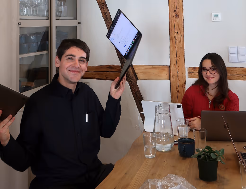 Zwei Personen sitzen an einem Holztisch mit Laptops, ein Mann lächelt und hält einen Laptop hoch, eine Frau mit Brille und rotem Oberteil lächelt.