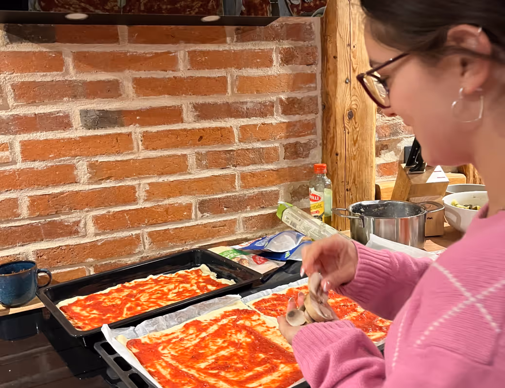 Person in einem rosa Pullover legt geschnittene Champignons auf rechteckigen Pizzateig mit Tomatensauce auf einer hölzernen Küchenarbeitsplatte.