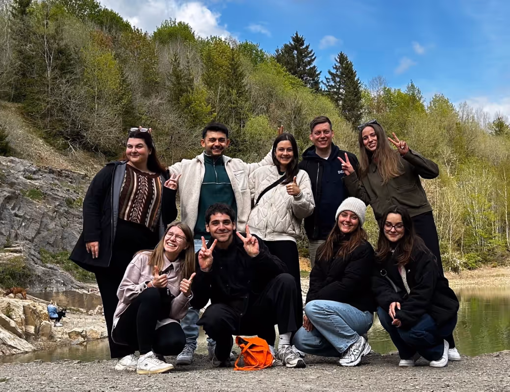 Gruppe von neun jungen Erwachsenen, die im Freien in der Nähe eines felsigen Bereichs und eines kleinen Gewässers posieren, mit Bäumen und blauem Himmel im Hintergrund.