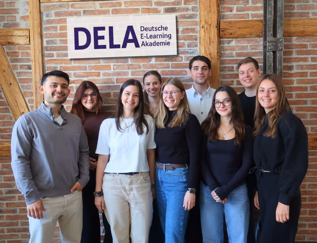 Gruppe von neun lächelnden jungen Erwachsenen, die vor einer Backsteinwand mit einem Schild der DELA Deutsche E-Learning Akademie stehen.