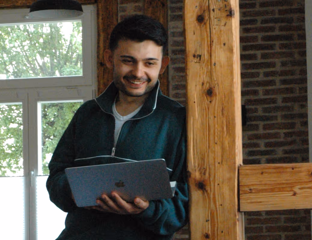 Lächelnder Mann, der sich gegen einen Holzbalken lehnt und einen offenen Laptop hält, nahe einem Fenster mit Grün draußen.