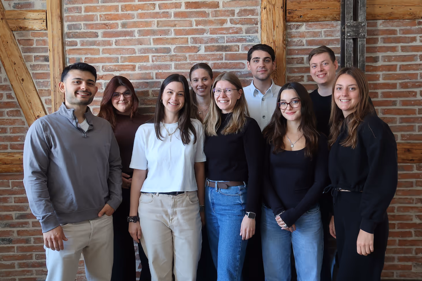 Gruppe von neun jungen Leuten, die vor einer Backsteinwand mit Holzbalken stehen und lächeln.