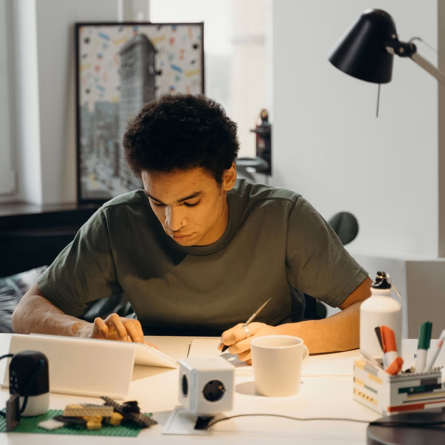 Junger Mann mit grünem T-Shirt sitzt an einem Tisch, zeichnet konzentriert auf Papier, mit Tablet, Tasse und Bastelmaterialien um ihn herum.