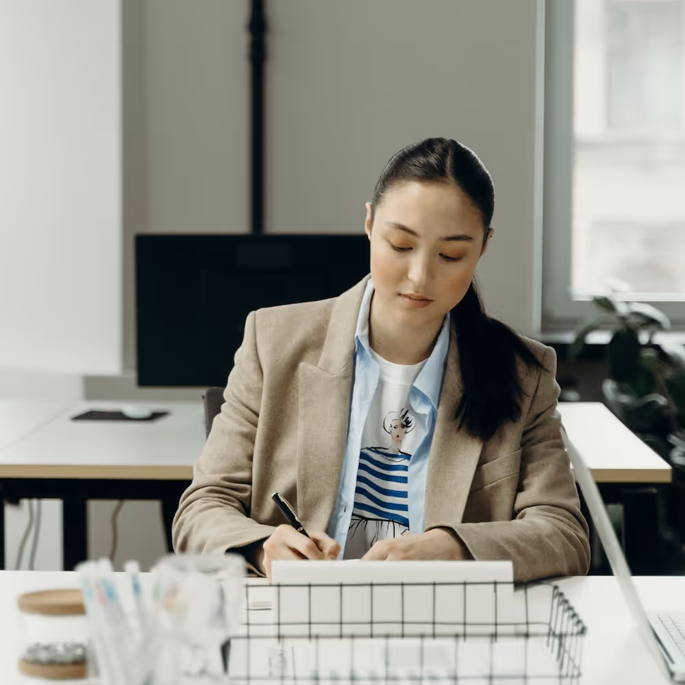 Person in beigem Blazer hält einen schwarzen Stift und zeigt auf ein Notizbuch, das auf einem weissen Schreibtisch liegt.