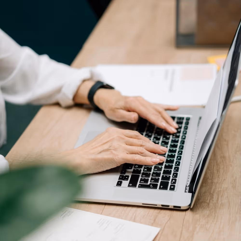 Ältere Person mit weissem Hemd tippt auf einem Laptop, auf einem Holztisch mit Unterlagen und Dokumenten.
