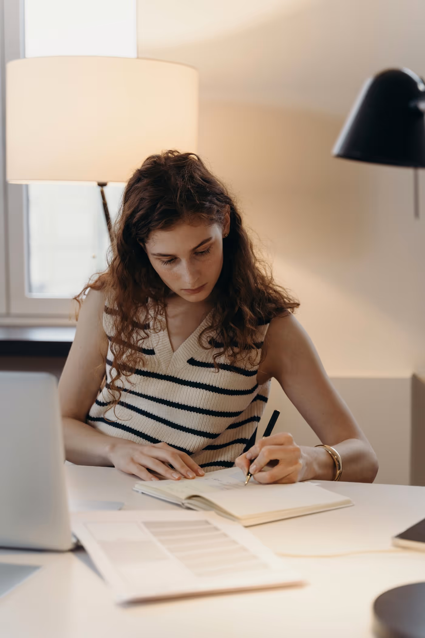 Junge Frau mit langen, lockigen Haaren und ärmellosem Oberteil schreibt in ein Notizbuch, während sie an einem Schreibtisch mit Laptop und Papieren sitzt.