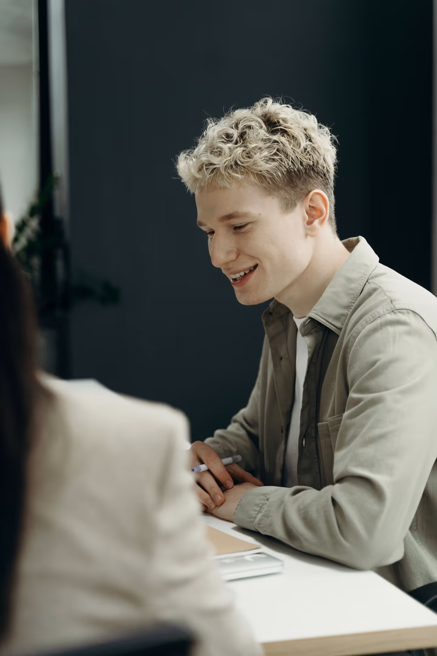 Junger Mann mit blond gelockten Haaren sitzt an einem Schreibtisch und lächelt während eines Gesprächs.