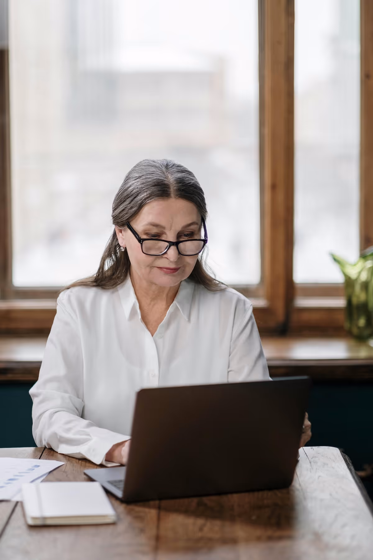 Ältere Frau mit Brille arbeitet an einem Laptop an einem Holztisch, mit einem grossen Fenster im Hintergrund.
