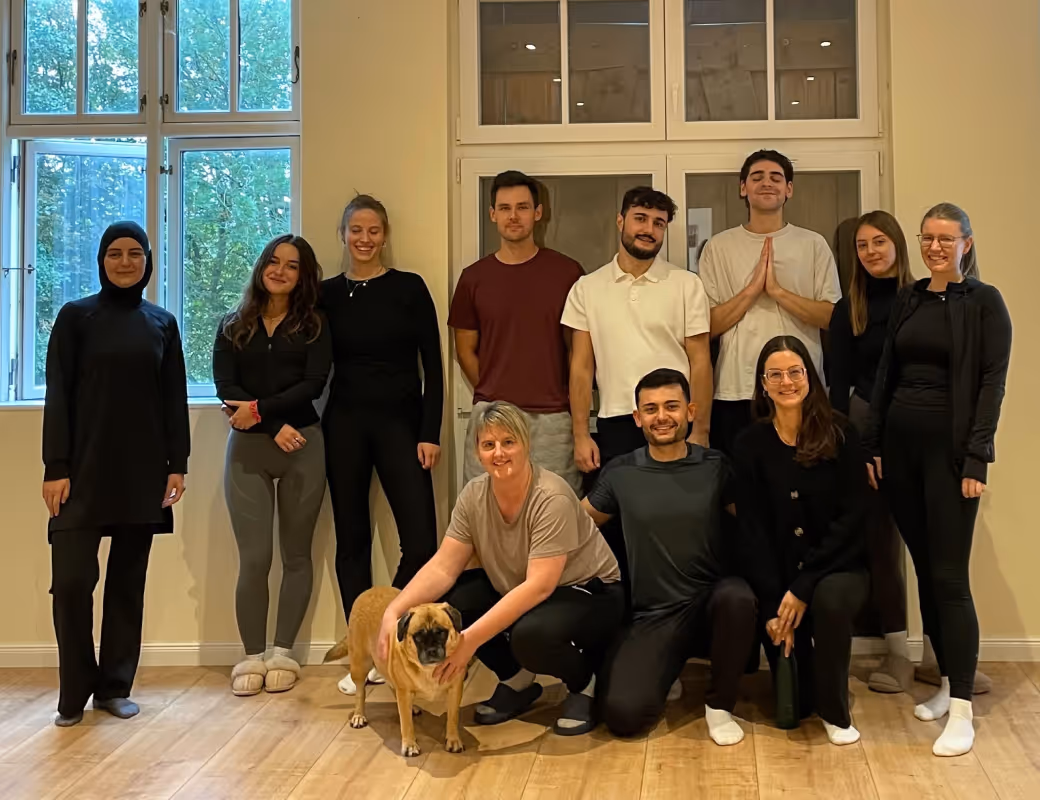 Gruppe von elf Personen, die drinnen auf einem Holzboden posieren, wobei eine Person hockt und einen hellbraunen Hund vorne hält.