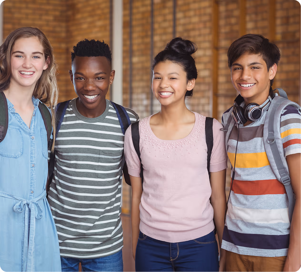 **Alt text:** “Four smiling students standing together indoors with backpacks, posing in a school hallway with a brick wall background.”