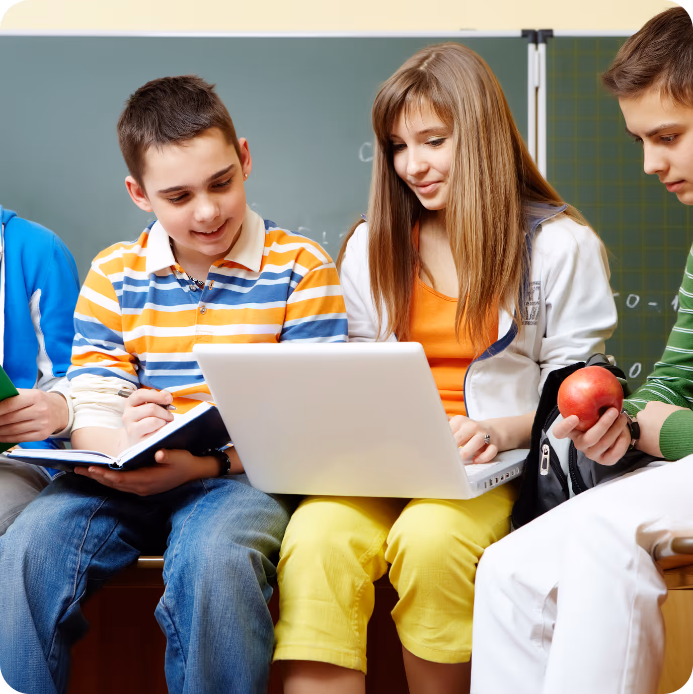 This image shows a group of students sitting together in a classroom, collaborating around a laptop. The central focus is on a young man in a colorful striped shirt and a young woman in an orange top with yellow pants, both looking at the laptop screen. Other students are visible on either side, creating a scene of engaged, collaborative learning in front of a chalkboard.