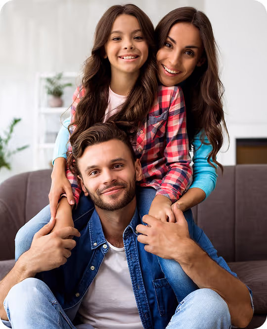 A warm family portrait showing a mother with two children - a teenage girl and a younger boy - all smiling at the camera in a bright, modern home setting with plants visible in the background. The mother wears a plaid shirt, the girl wears white, and the boy is in the foreground, creating an intimate, supportive family scene.