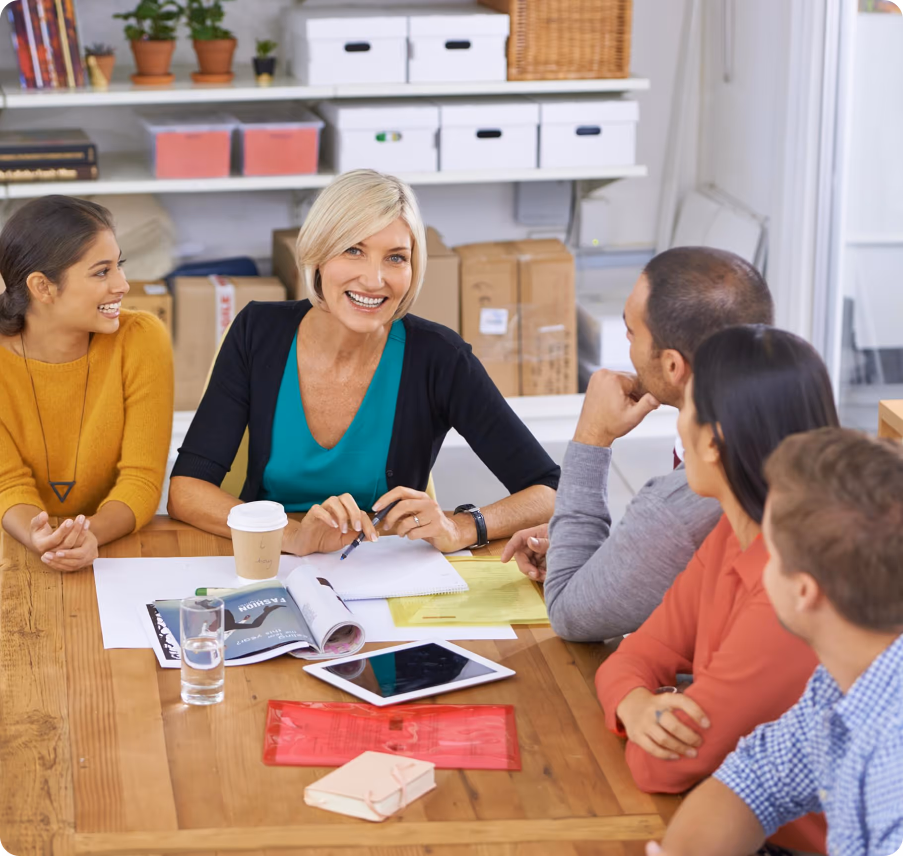 A blonde woman in a black cardigan and green top smiles at the camera while sitting at a wooden table with four other people in a meeting setting. The table is covered with papers, a tablet, coffee cups, and a glass of water, and shelves with white storage boxes are visible in the background.
