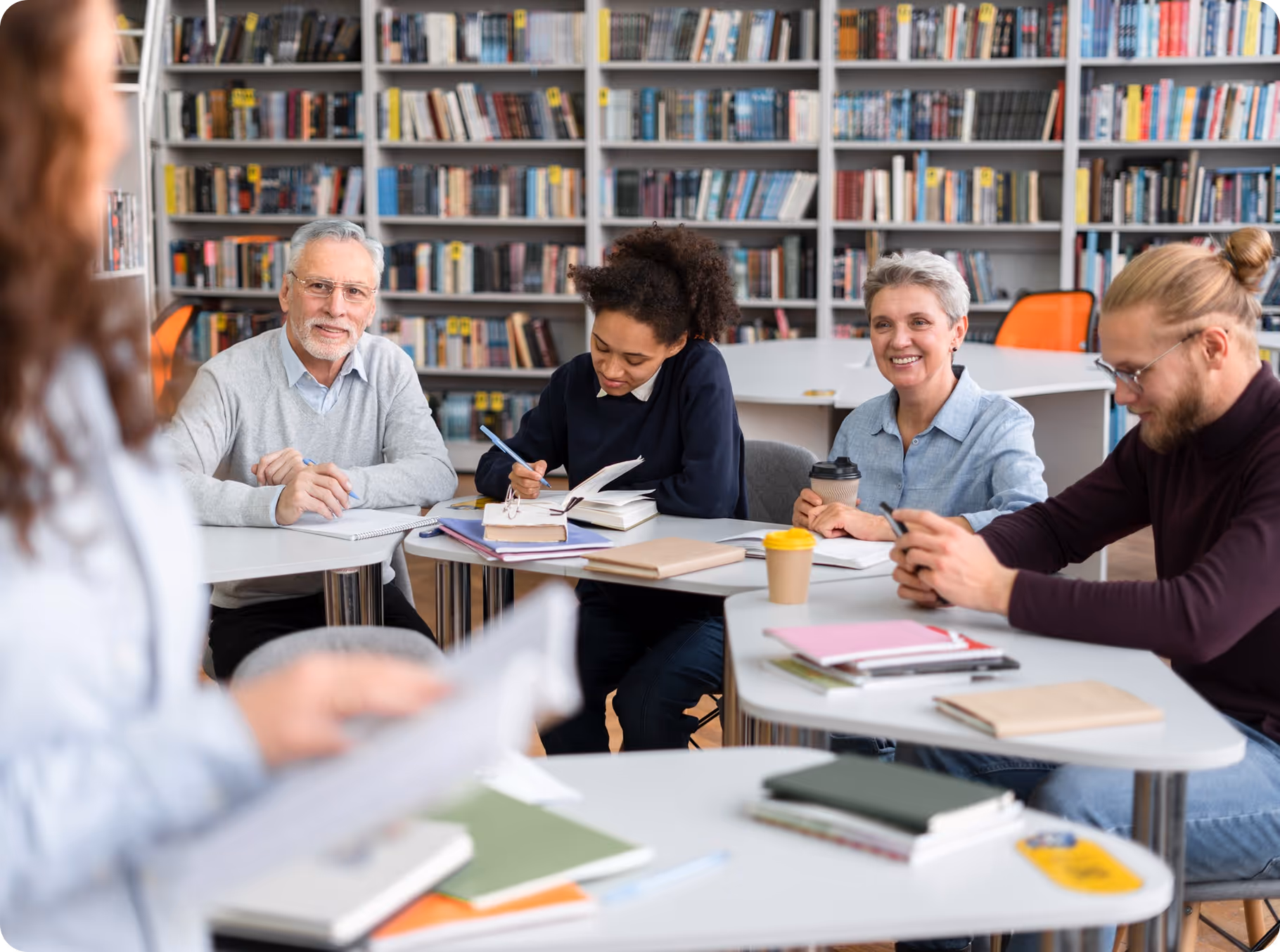 "Diverse group of students studying together at tables in library with extensive bookshelf backdrop, taking notes and discussing with coffee cups and notebooks scattered across workspace