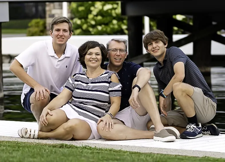 This image shows a family portrait of four people smiling at the camera against a gold sequined or glittery background. In the back row are two young men wearing light blue shirts, and in the front row are an older man with glasses wearing a blue button-down shirt and a woman wearing a navy and white patterned dress. Everyone is smiling warmly, creating a cheerful family photo.