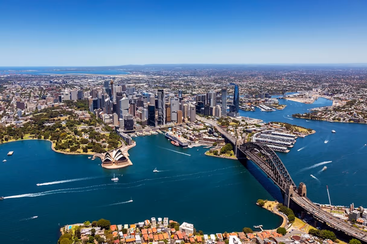 Aerial view of Sydney Harbour with the Sydney Opera House, Sydney Harbour Bridge, and city skyline under a clear blue sky.