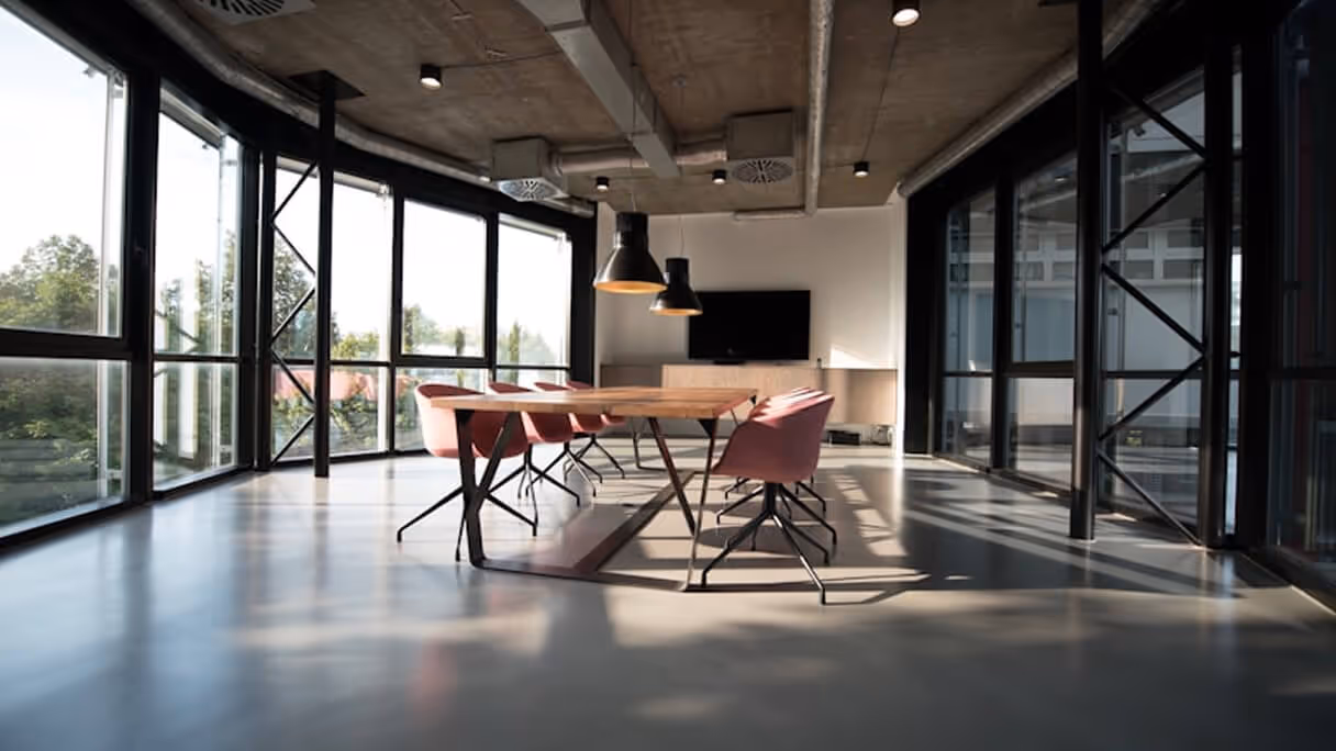 Modern conference room with large wooden table, pink chairs, pendant lights, and floor-to-ceiling windows.