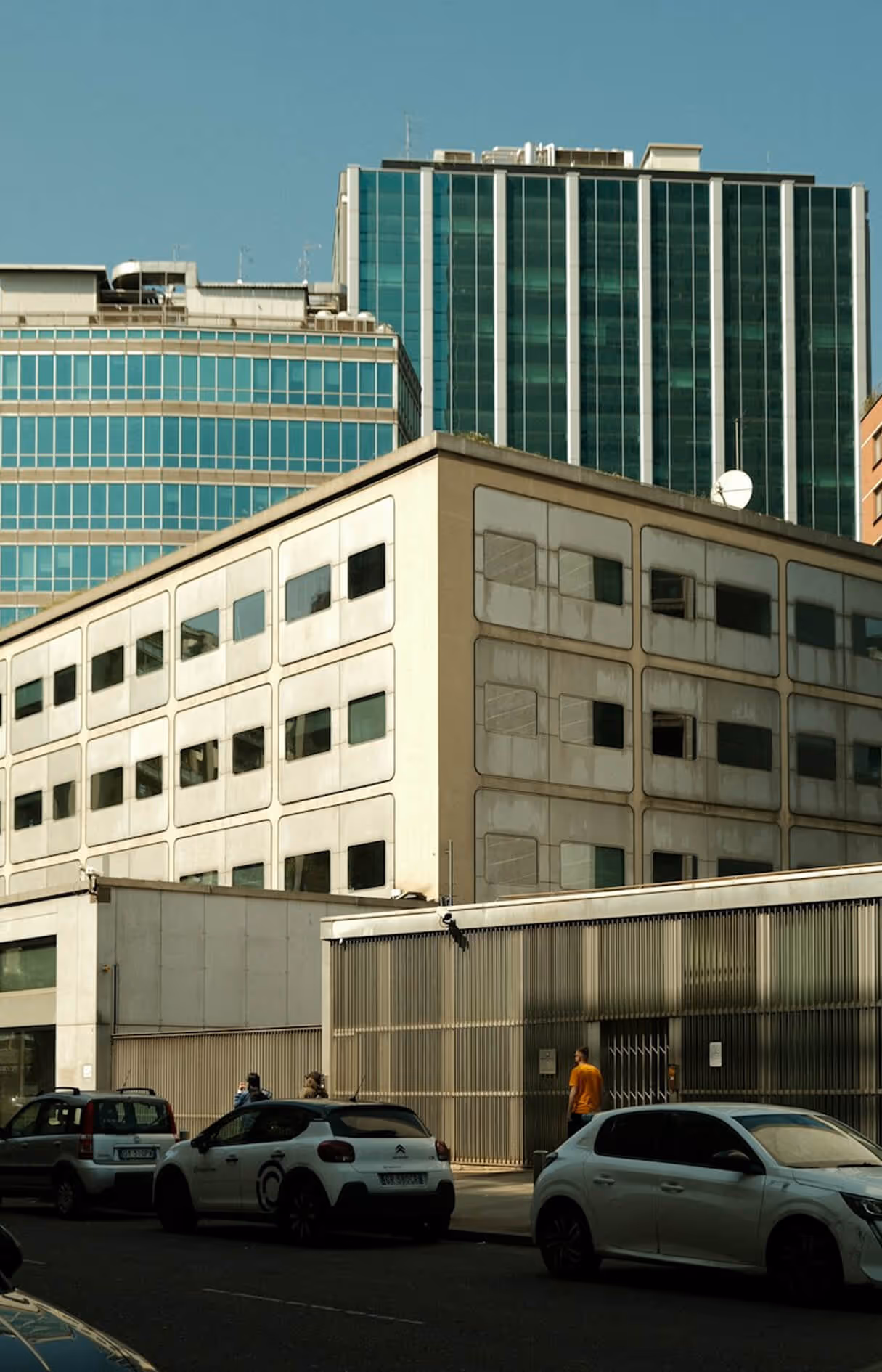 Urban street scene with parked cars and a person walking beside modern office buildings with tinted windows.