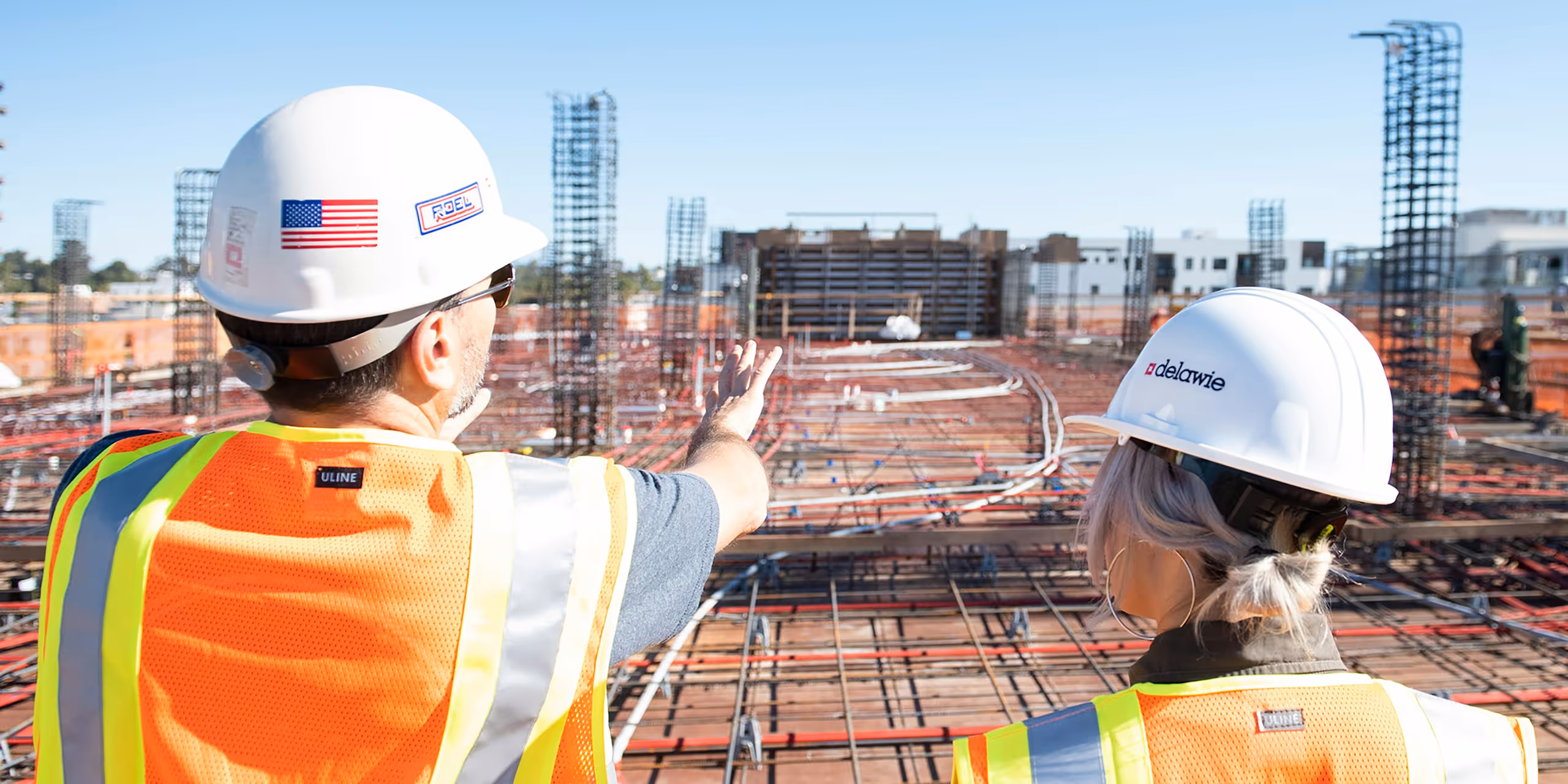 Two construction workers wearing white helmets and orange safety vests overlooking a building site with steel reinforcements under a clear blue sky.