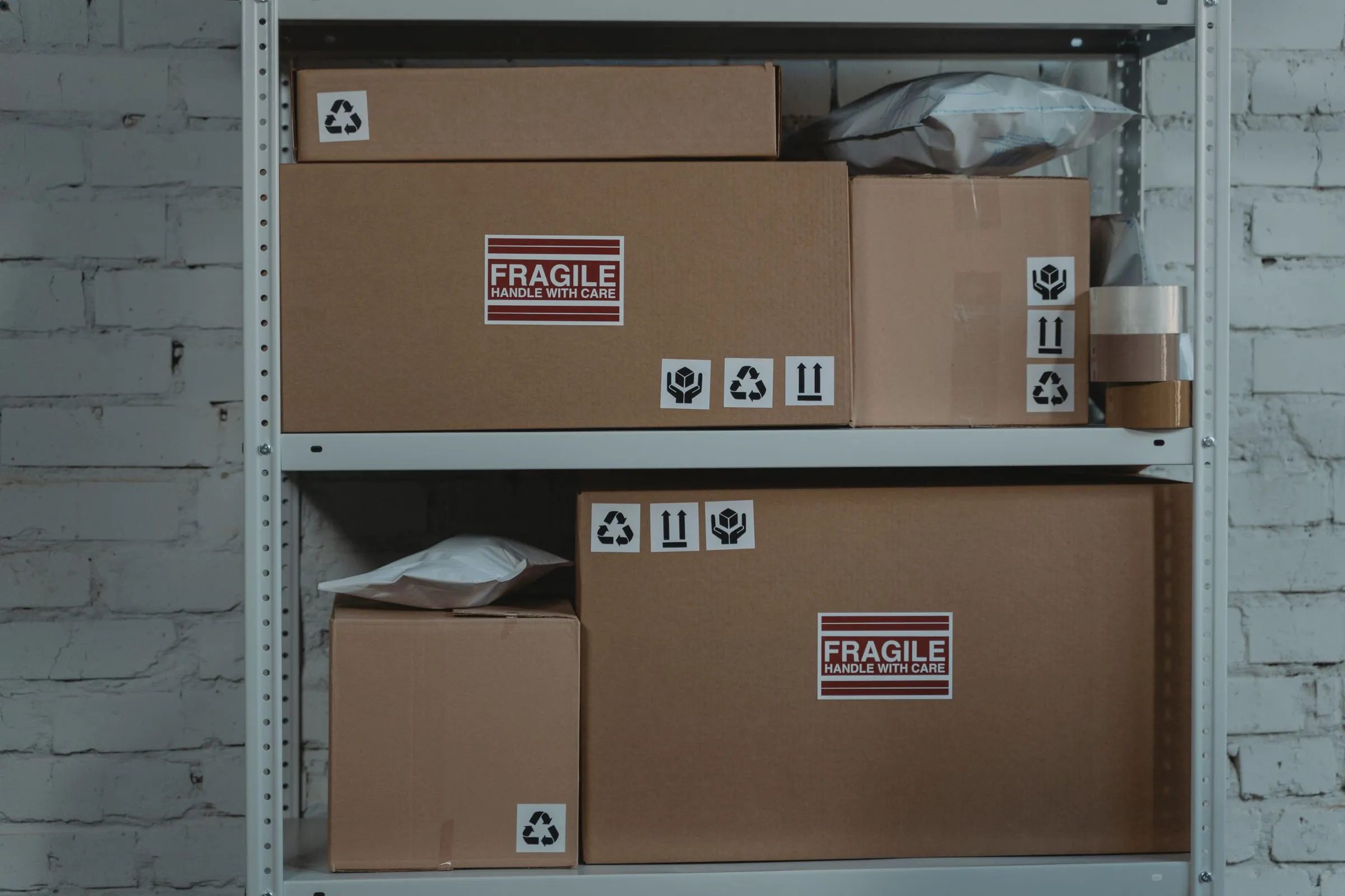 Metal shelving unit against a white brick wall holding stacked cardboard boxes and padded envelopes, some with 'fragile handle with care' labels and recycling, handle with care, and upright arrow symbols.