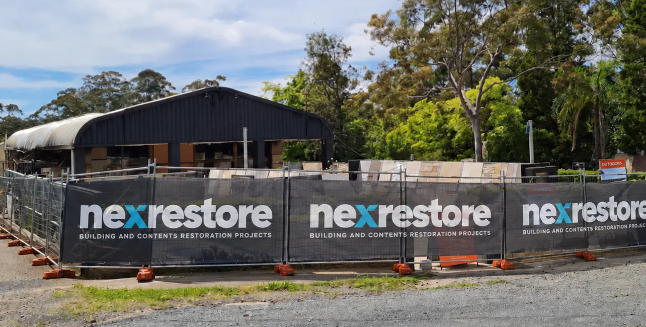 Construction site fenced off with black banners displaying the logo and text 'nexrestore BUILDING AND CONTENTS RESTORATION PROJECTS' repeated three times, with a partially covered building and trees in the background.