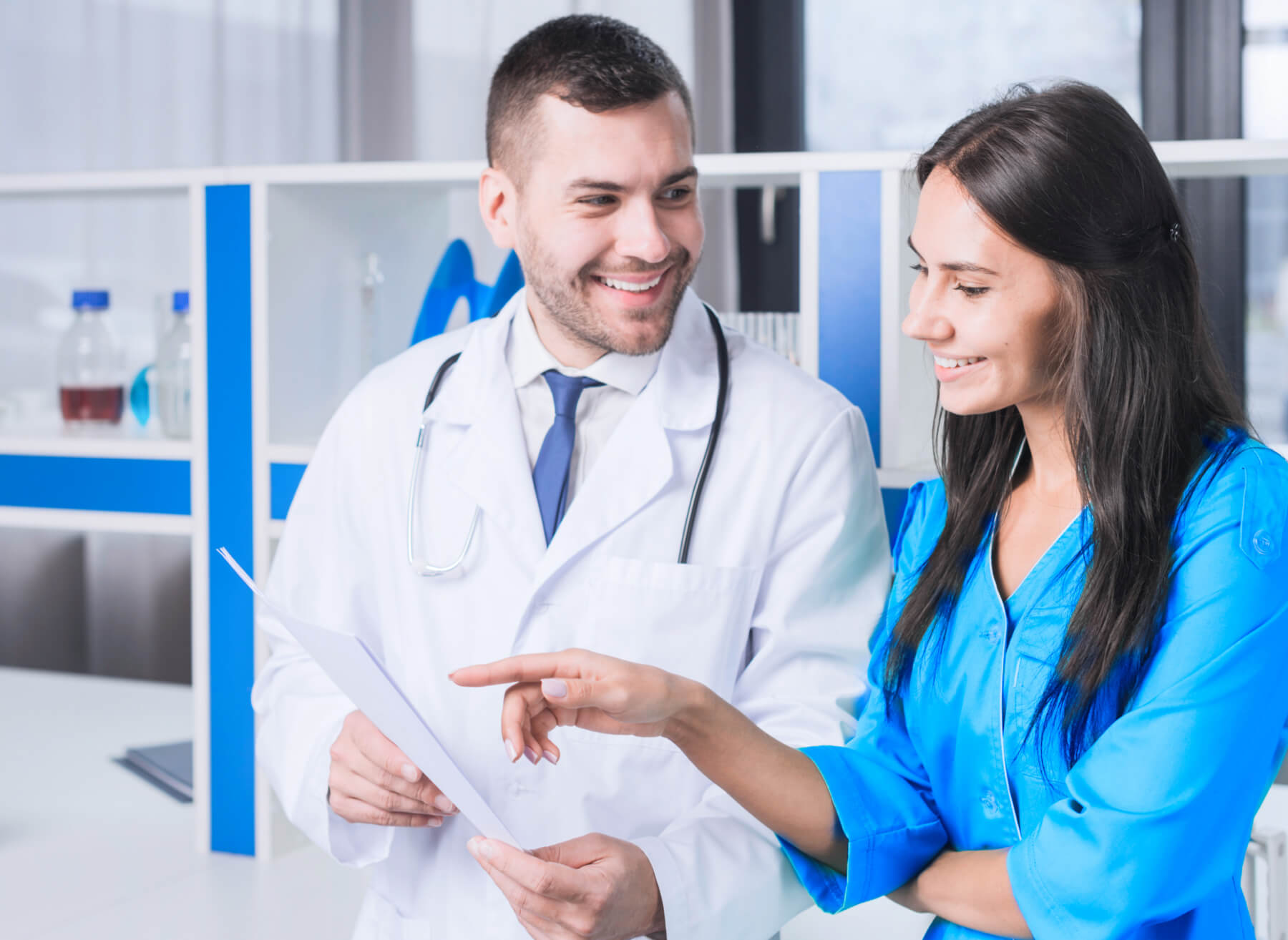 Smiling doctor in white coat holding a clipboard while a woman in blue shirt points at it, both engaging in discussion inside a medical office.