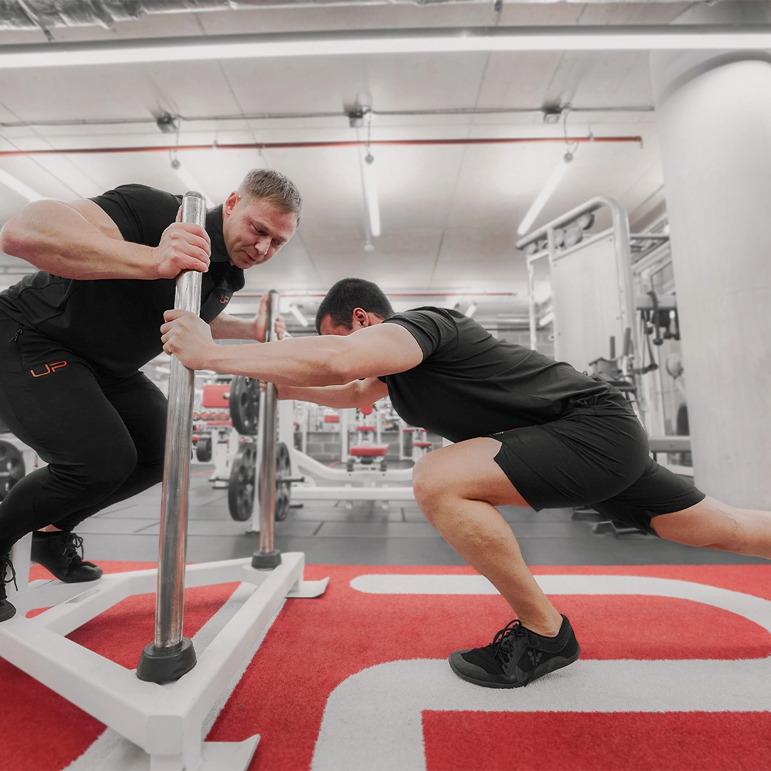 Trainer assisting a man stretching his legs while holding onto a metal sled in a gym.