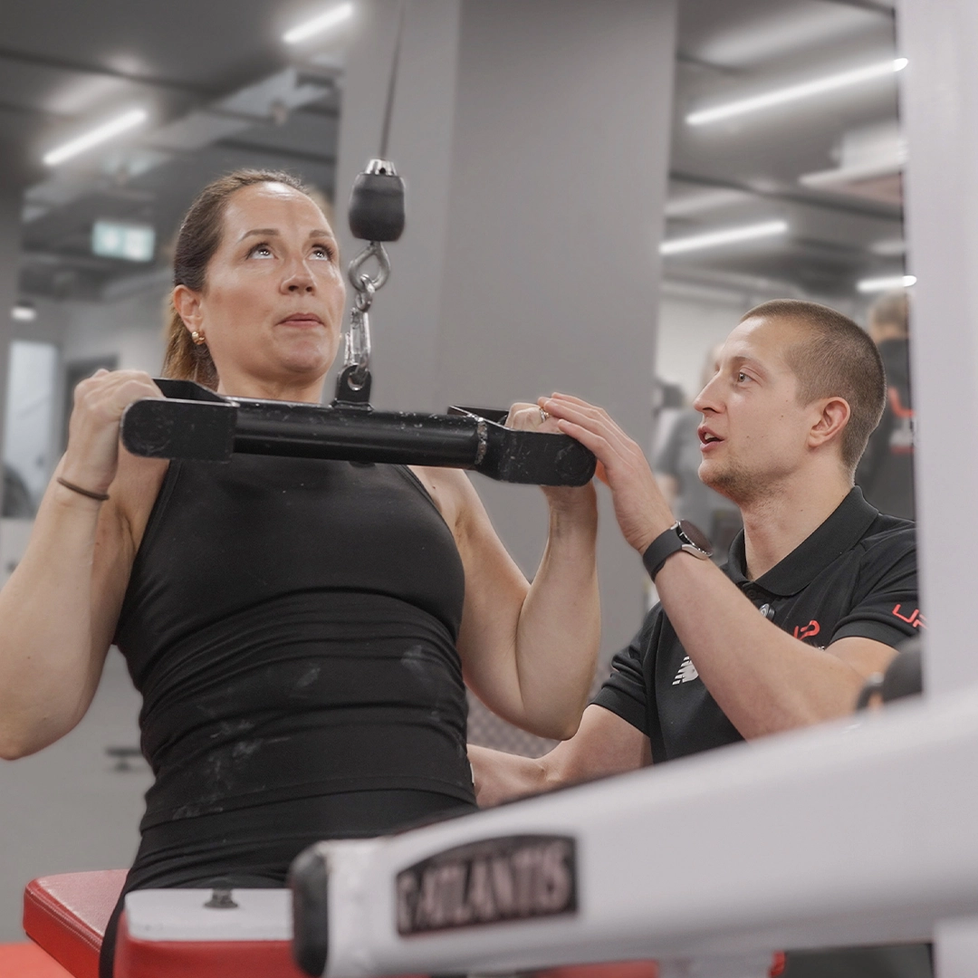 Woman performing seated lat pulldown exercise with trainer assisting in a gym.
