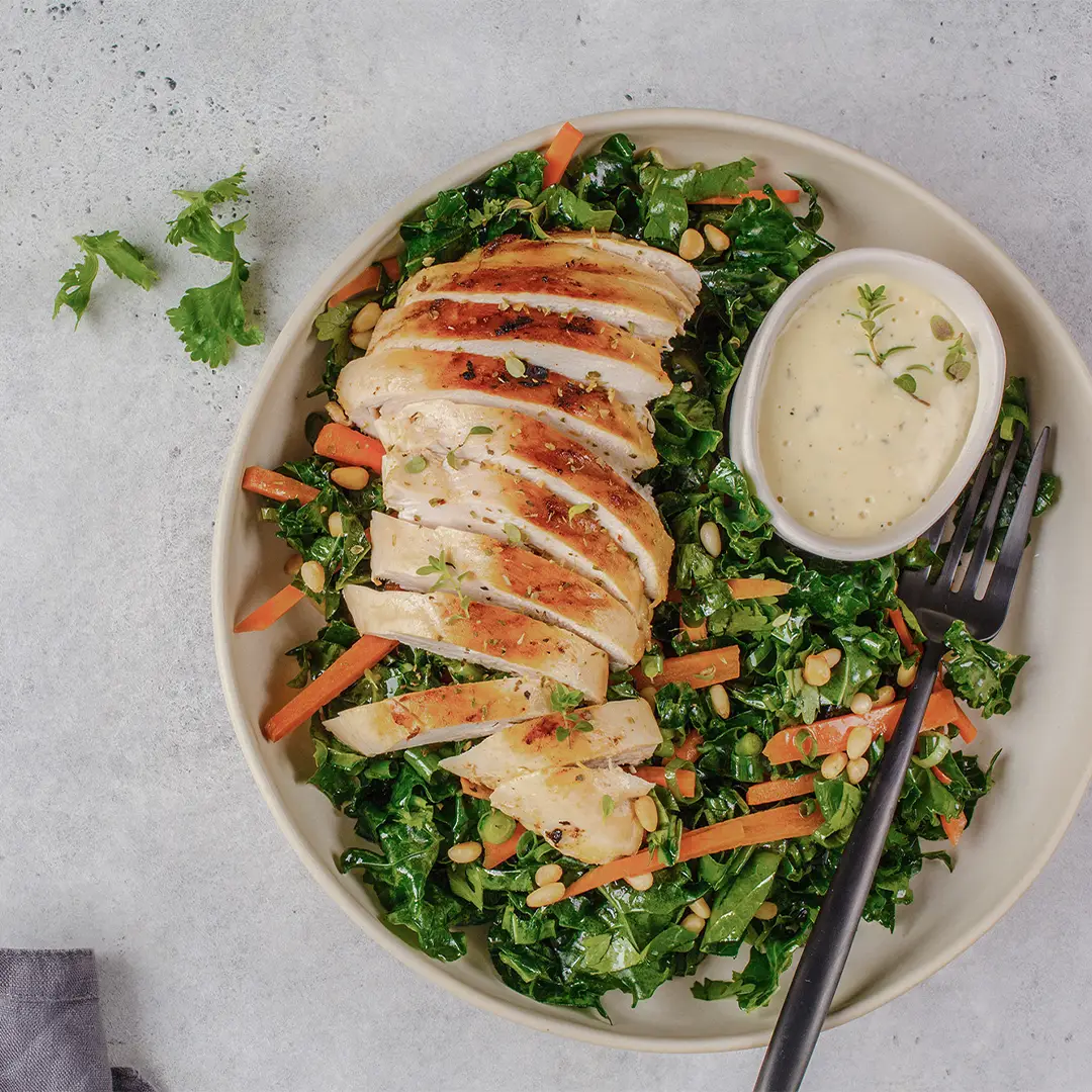 Sliced grilled chicken breast served on a bed of kale and carrot salad with pine nuts and a small bowl of creamy dressing, accompanied by a black fork.
