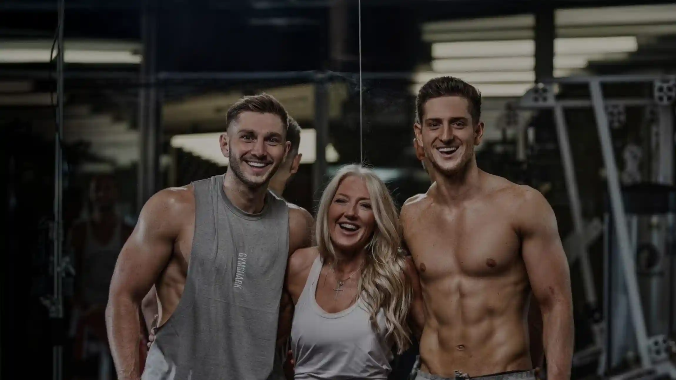 Two muscular men and a smiling woman standing closely together in a gym.
