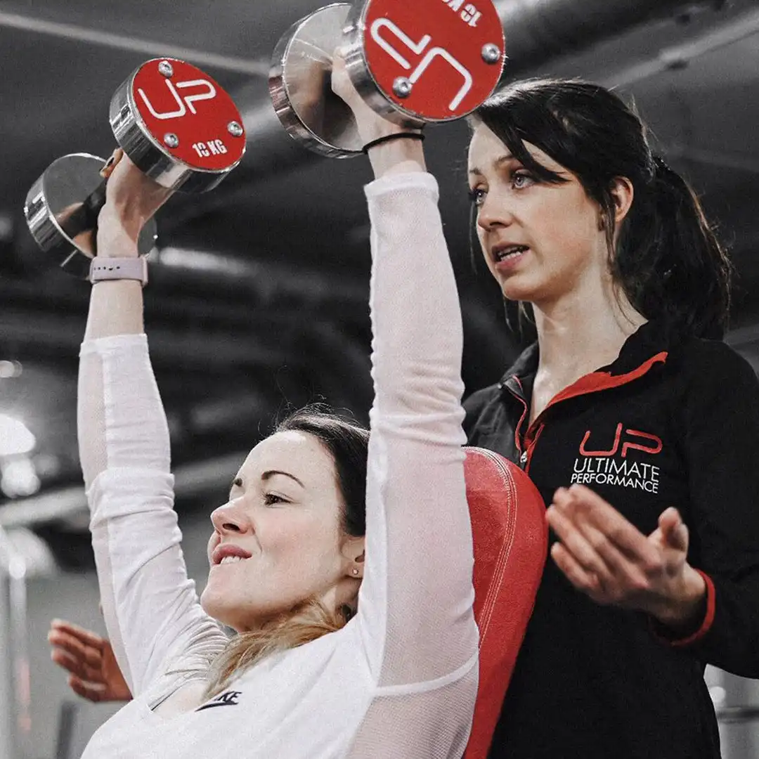 Woman lifting dumbbells overhead on a bench with a female trainer coaching her in a gym.