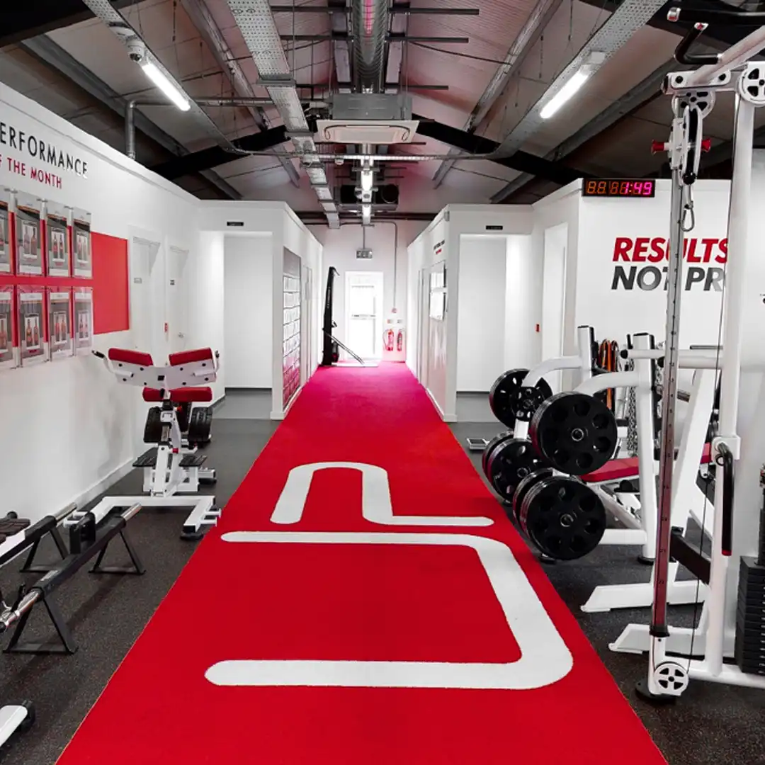 Modern gym interior with a red carpeted track, white workout machines, and performance achievement boards on the wall.