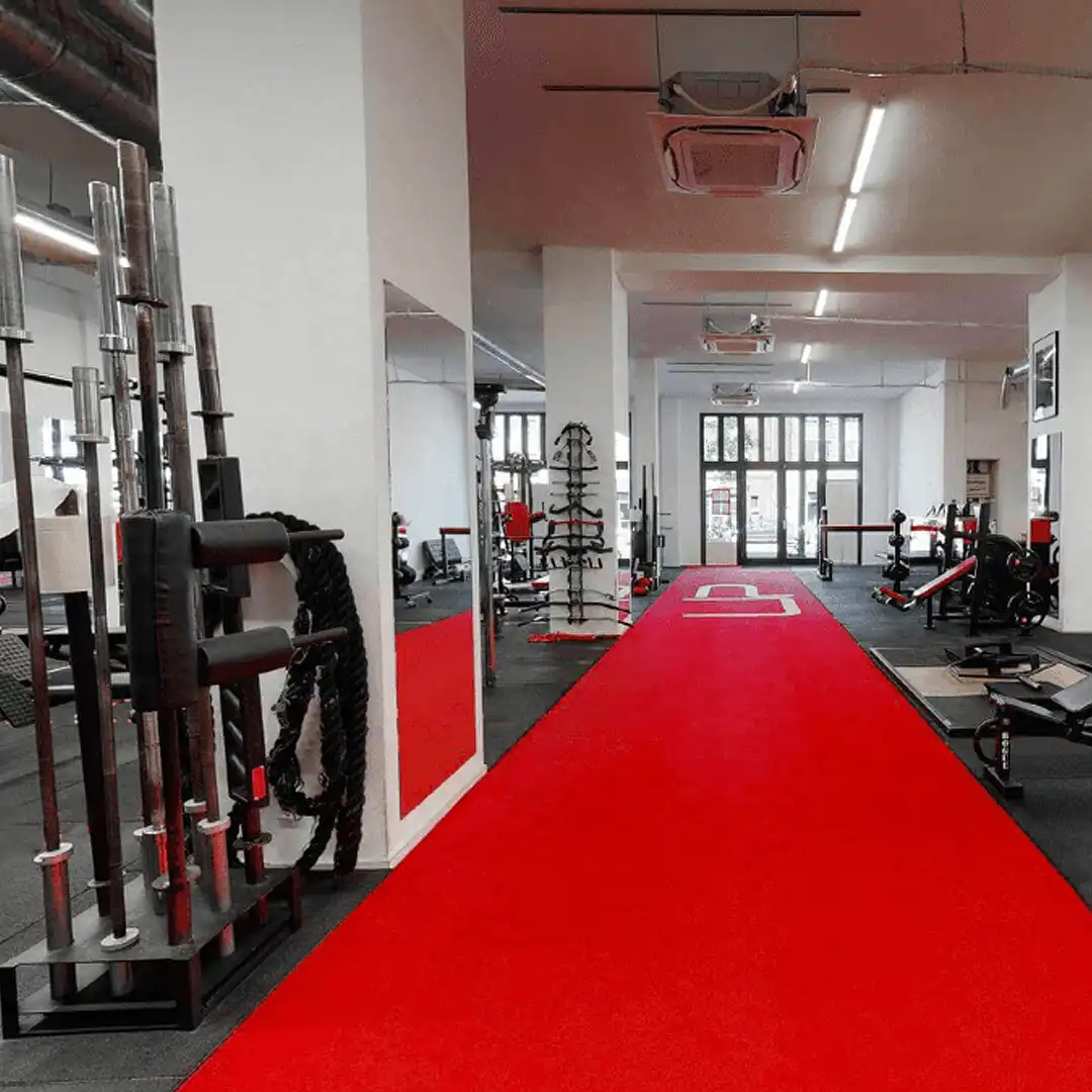 Modern gym interior with a red track carpet, weightlifting bars, benches, and large windows in the background.