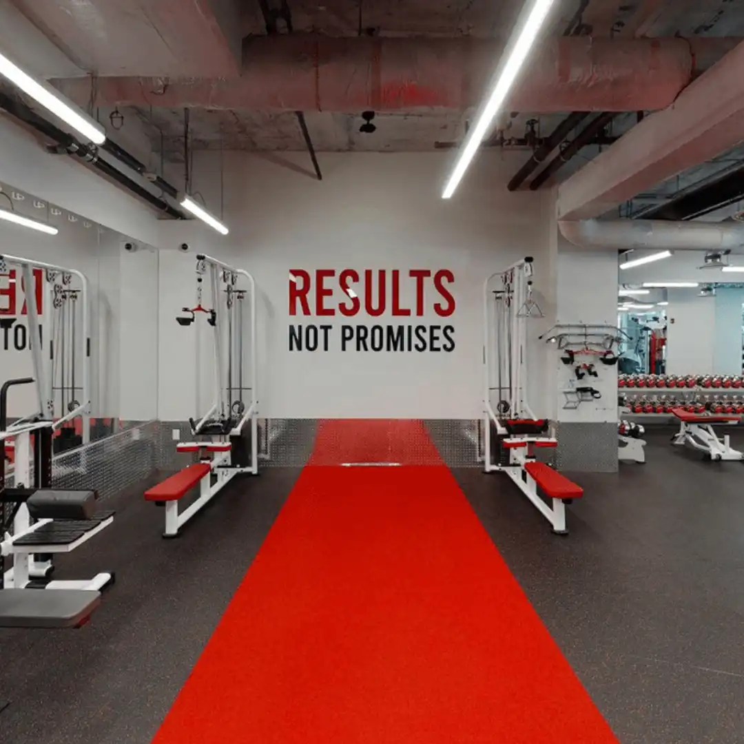 Interior of a gym with red and black workout equipment, a red floor track, and the phrase 'RESULTS NOT PROMISES' on a white wall.