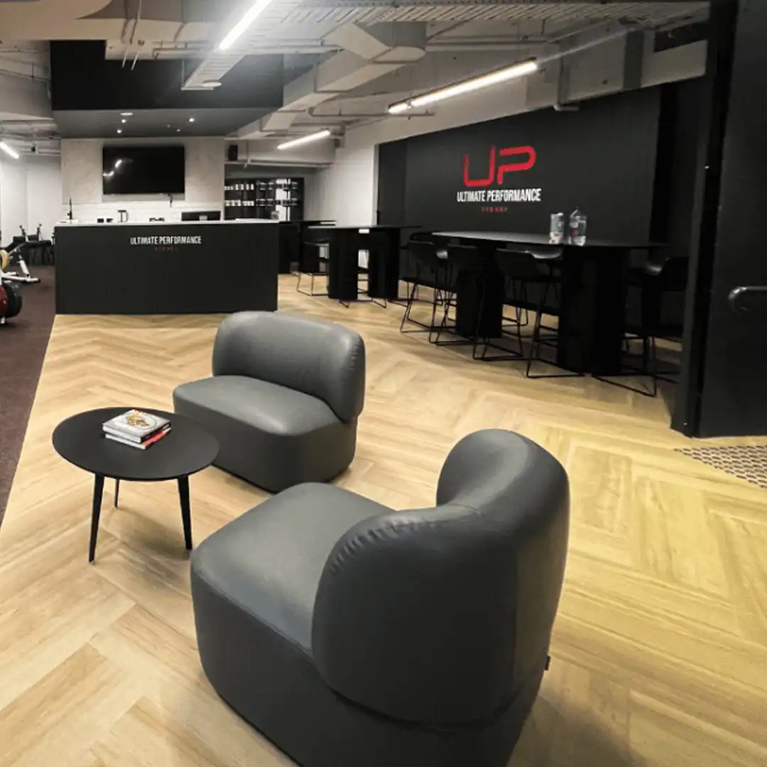 Modern gym lounge area with two gray armchairs, a small round black table, and a black reception desk with Ultimate Performance branding.