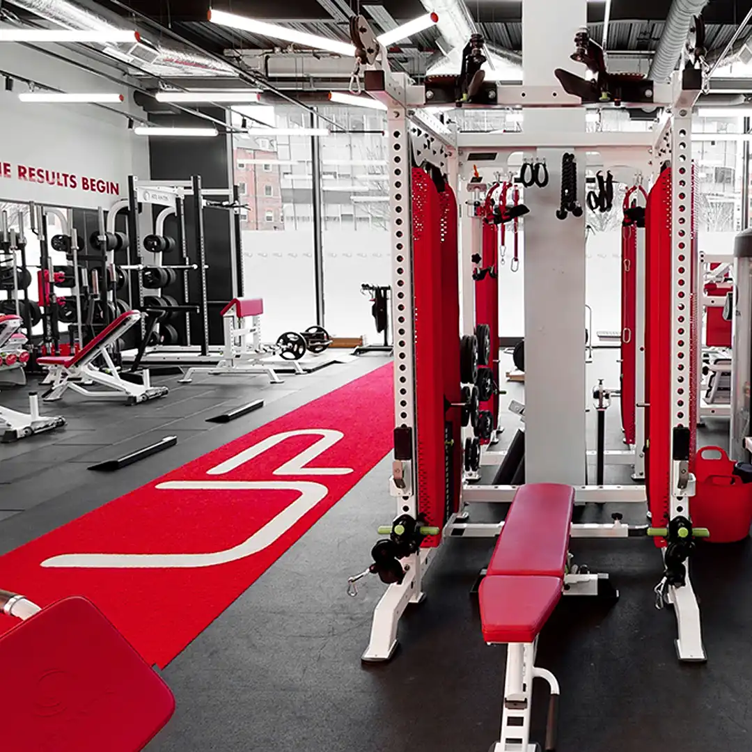 Modern gym interior with weightlifting equipment, red and white benches, and a red carpet with a white logo.