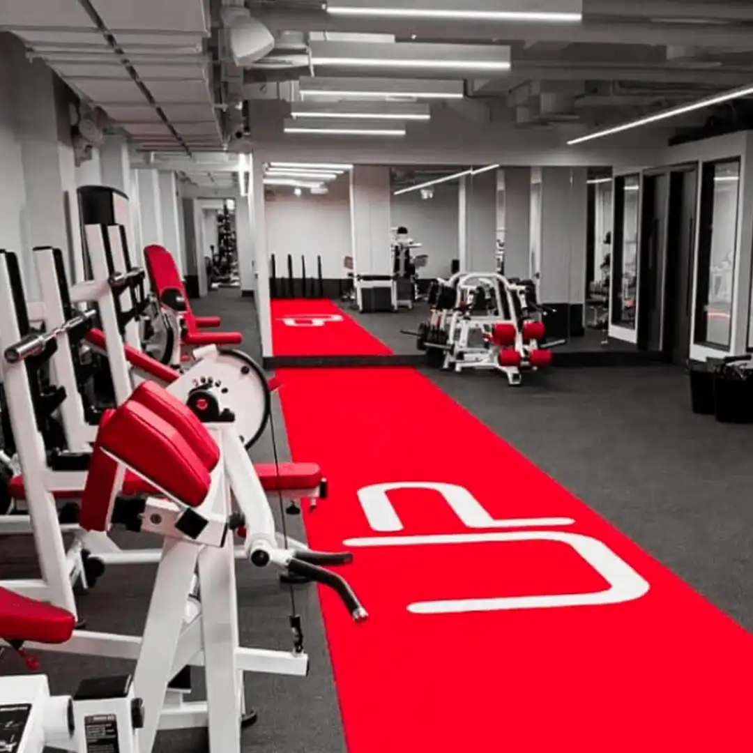 Modern gym interior with white and red workout machines and a red carpet with white logo in the center.