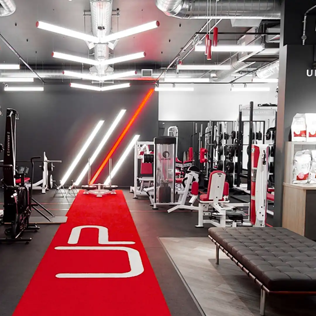Modern gym interior with red carpet pathway, various workout machines, and stylish lighting fixtures.