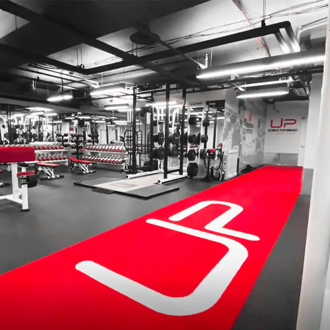 Modern gym interior with weightlifting racks, benches, dumbbells, and a red floor mat with large white UP logo.