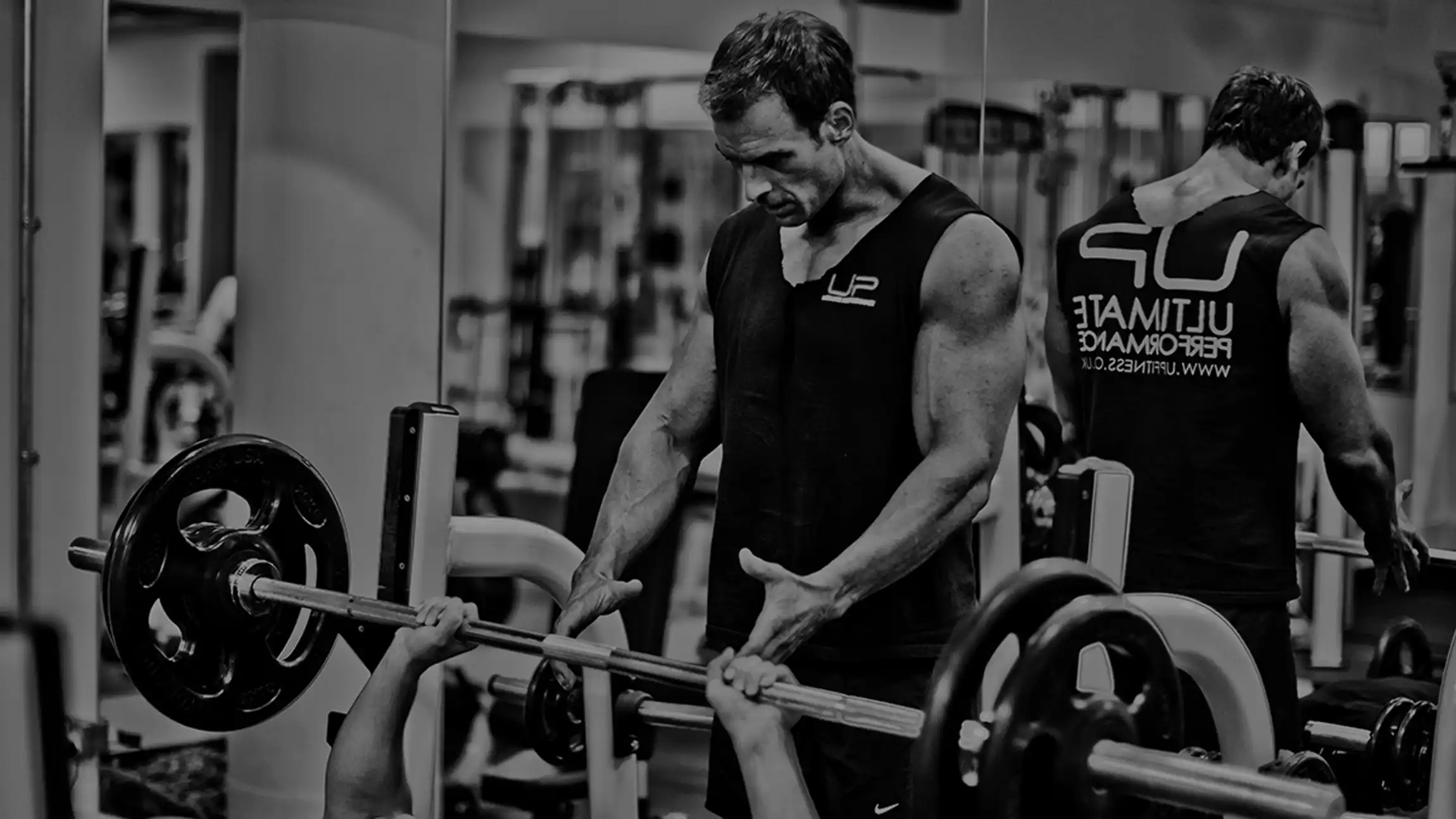 A muscular man in a sleeveless shirt spotting another person lifting a barbell in a gym.