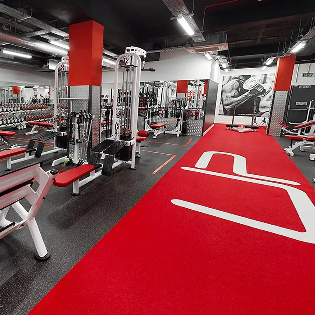 Modern gym interior with red and black flooring, weight machines, benches, and a large black-and-white photo of a muscular arm lifting a dumbbell.