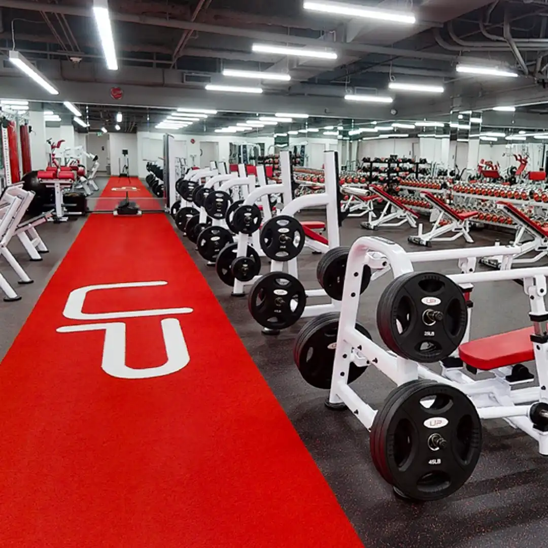 Spacious gym room with weightlifting benches, barbells, dumbbells, and a red carpet walkway featuring white CU lettering.