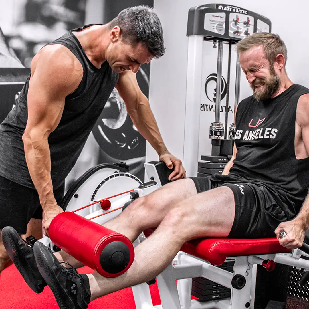 A muscular man assists another man who is straining while using a leg extension machine in a gym.