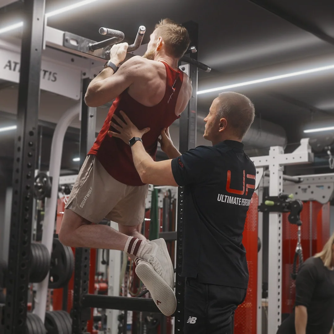 Trainer assisting a man doing pull-ups in a gym with weightlifting equipment in the background.